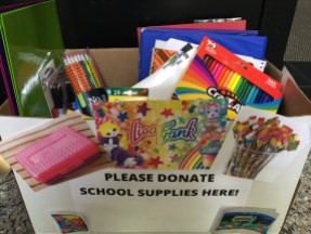A box full of folders, binders, pencils, and colored pencils. The box has a sign that says "Please donate school supplies here."